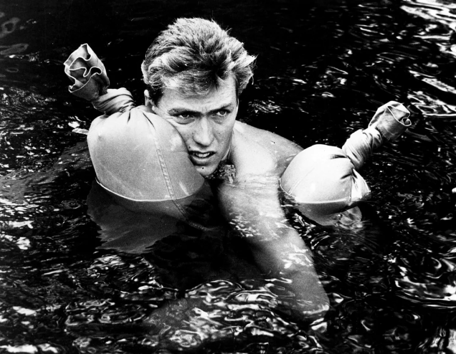 A man with wet hair floats in water wearing improvised water wings made from inflated objects tied at the ends, looking determined and focused.
