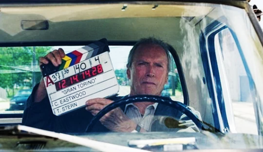 An older man sits behind the wheel of a car, holding a movie clapperboard labeled "Gran Torino" in front of him, viewed through the windshield.