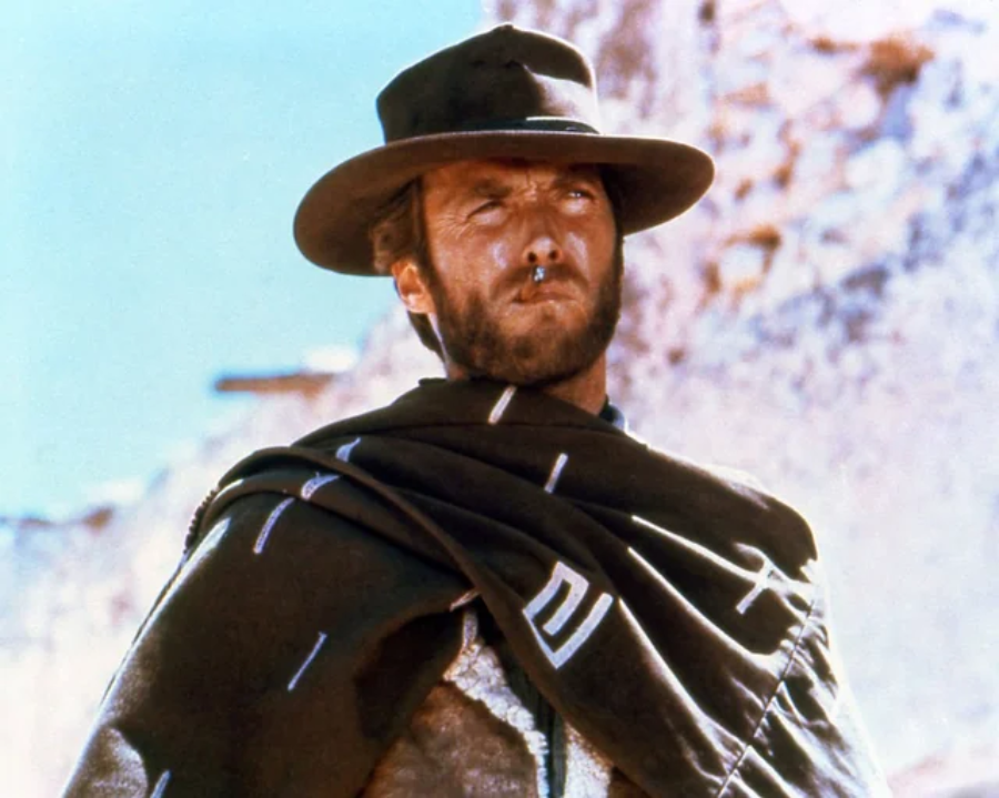A man wearing a wide-brimmed hat and dark poncho stands outdoors in a western setting, with a scruffy beard and a serious expression, holding a cigar in his mouth. The background is bright and sandy.