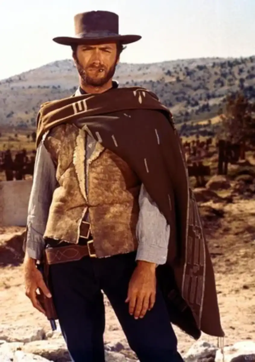 A man in a cowboy outfit stands outdoors, wearing a wide-brimmed hat, poncho, and vest. He has a scruffy beard, a cigar in his mouth, and rests his hand near a holstered gun. The background shows a dry, open landscape.