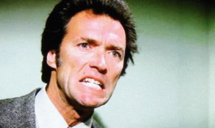A man with dark hair wearing a suit jacket and tie appears angry, baring his teeth and furrowing his brow against a greenish background.