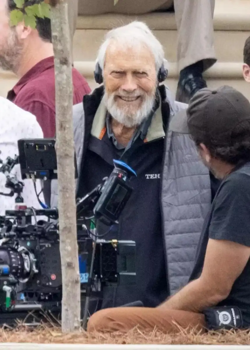 An older man with a white beard is smiling while wearing headphones on a film set, standing next to a camera and talking with another crew member outdoors.