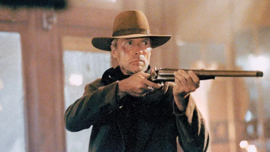 A man in a brown hat and coat holds a double-barreled shotgun, aiming intently. The background appears to be indoors with warm lighting and blurred windows.