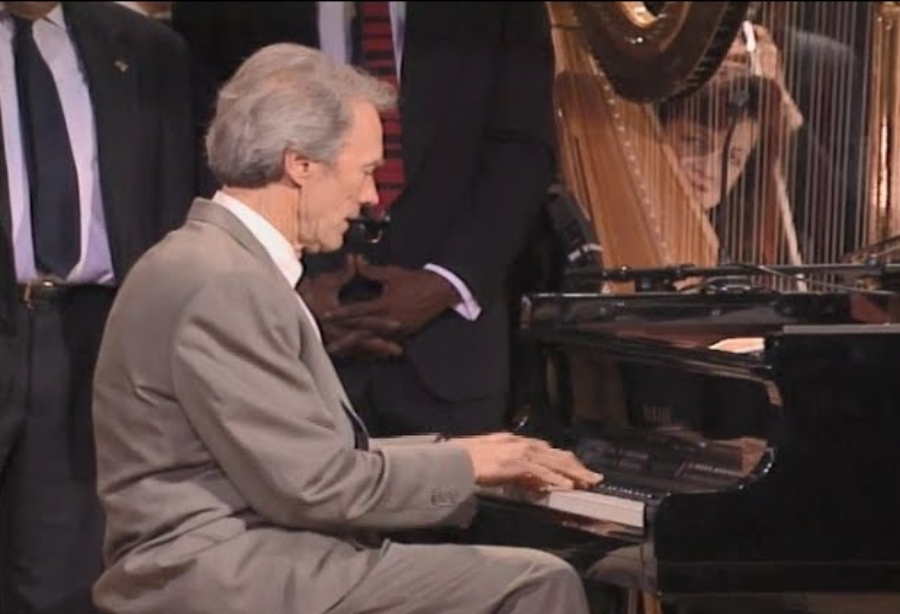 An older man in a gray suit plays a grand piano, with several people standing behind him and a harp visible to the right. A woman smiles in the background, reflected in the harp's strings.
