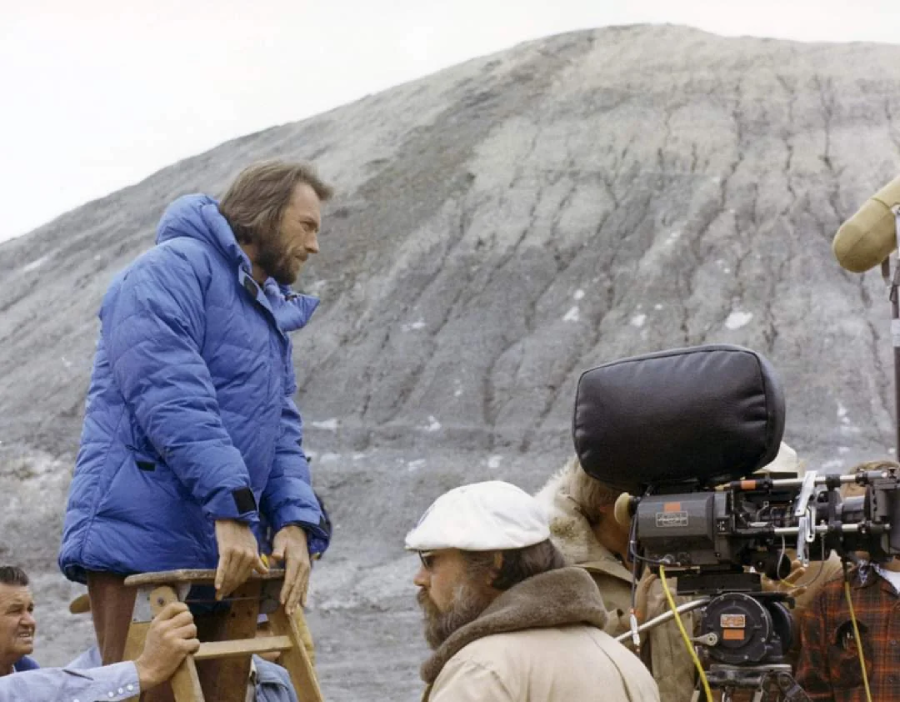 A man in a blue coat stands on a ladder near a rocky hillside, while crew members, a film camera, and a microphone are visible in the foreground during an outdoor film shoot.