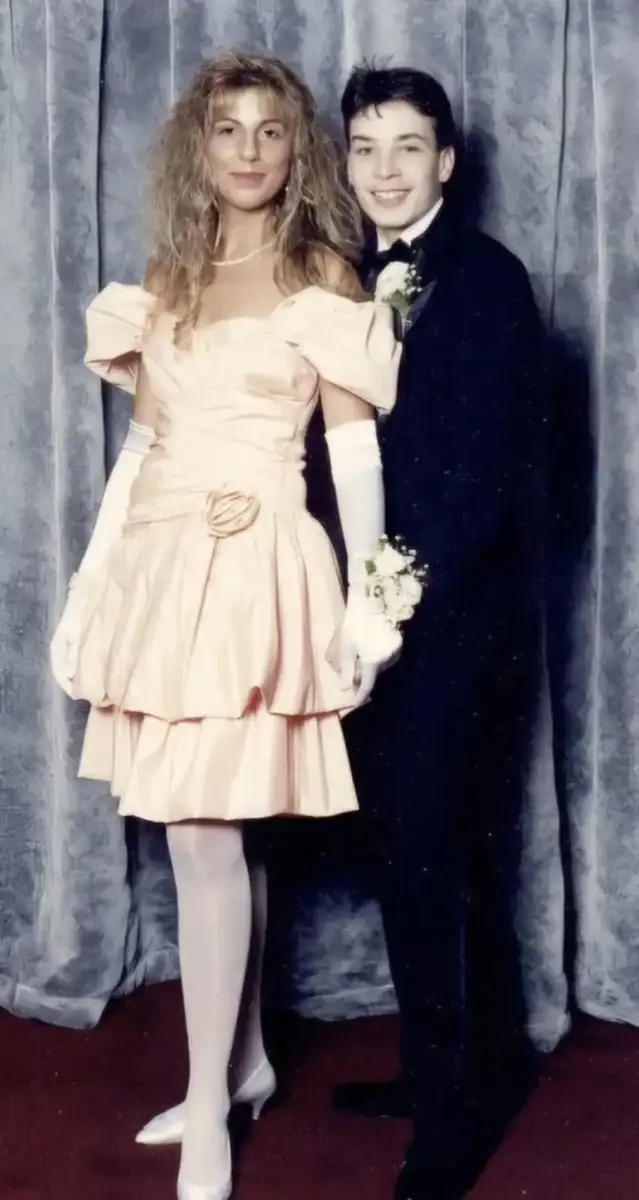 A young woman in a pink ruffled dress and white gloves stands beside a young man in a black tuxedo. They both smile in front of a gray curtain backdrop, dressed formally for an event, possibly prom.