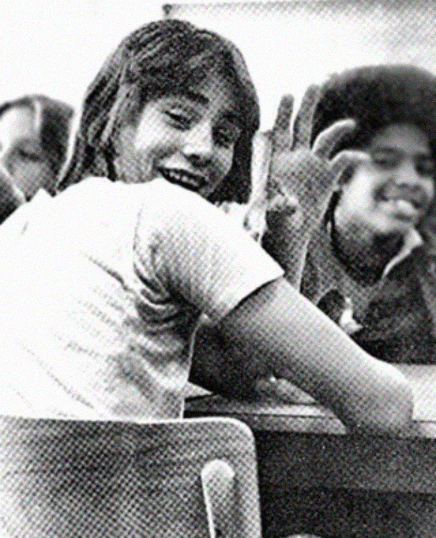 Two teenagers sit at desks, smiling and facing the camera. One waves with two fingers raised while the other looks over their shoulder, grinning. The image is black and white with a grainy texture.