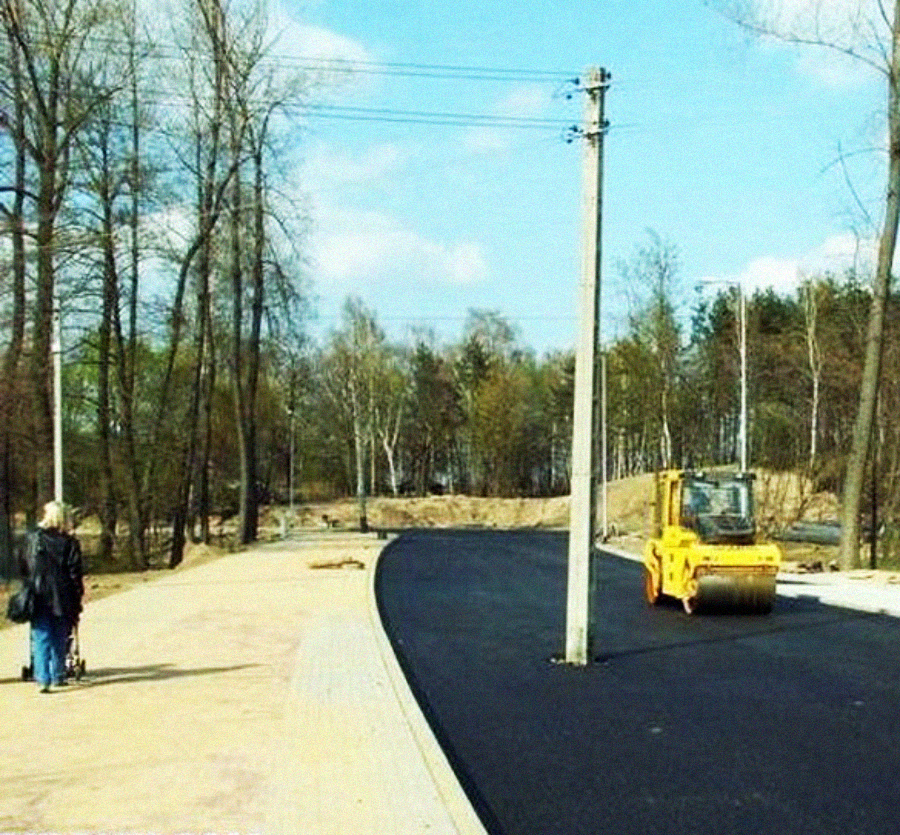 A paved road with a large utility pole awkwardly standing in the middle, while a road roller is nearby. Two people walk on a sidewalk beside the road, surrounded by trees and a clear sky.