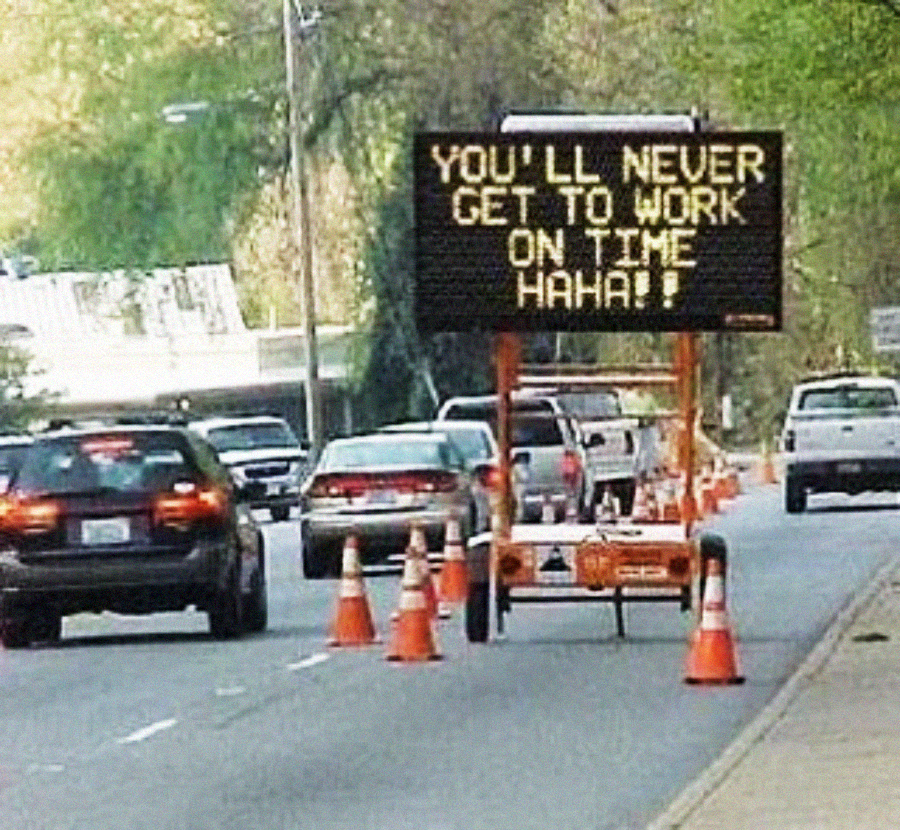 A portable electronic road sign on a street reads, "YOU'LL NEVER GET TO WORK ON TIME HAHA!!" with traffic cones and cars driving past in the background.