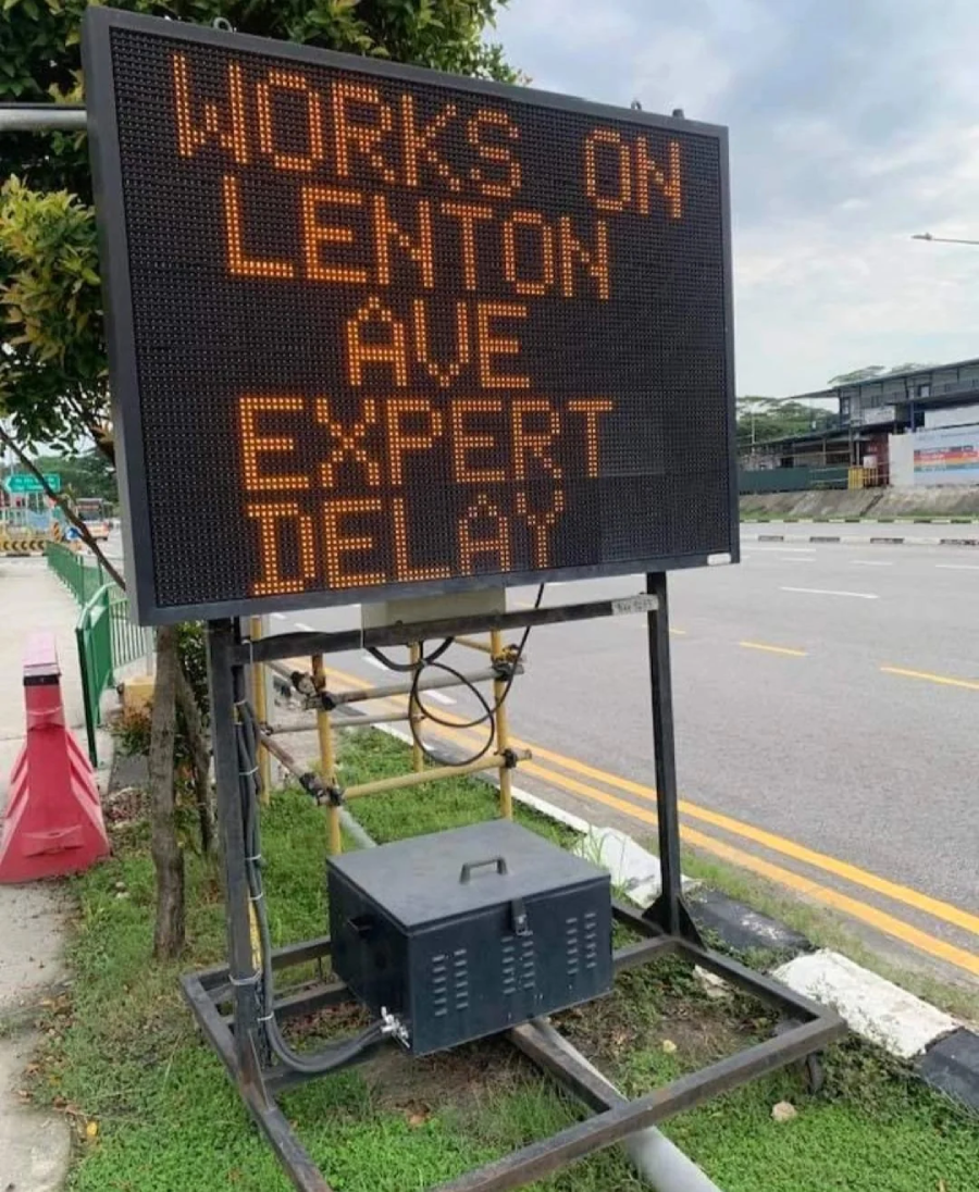 A roadside electronic sign reads "WORKS ON LENTON AVE EXPECT DELAY," though "EXPECT DELAY" is misspelled as "EXPERT DELAY." The sign is set up next to a street with minimal traffic.