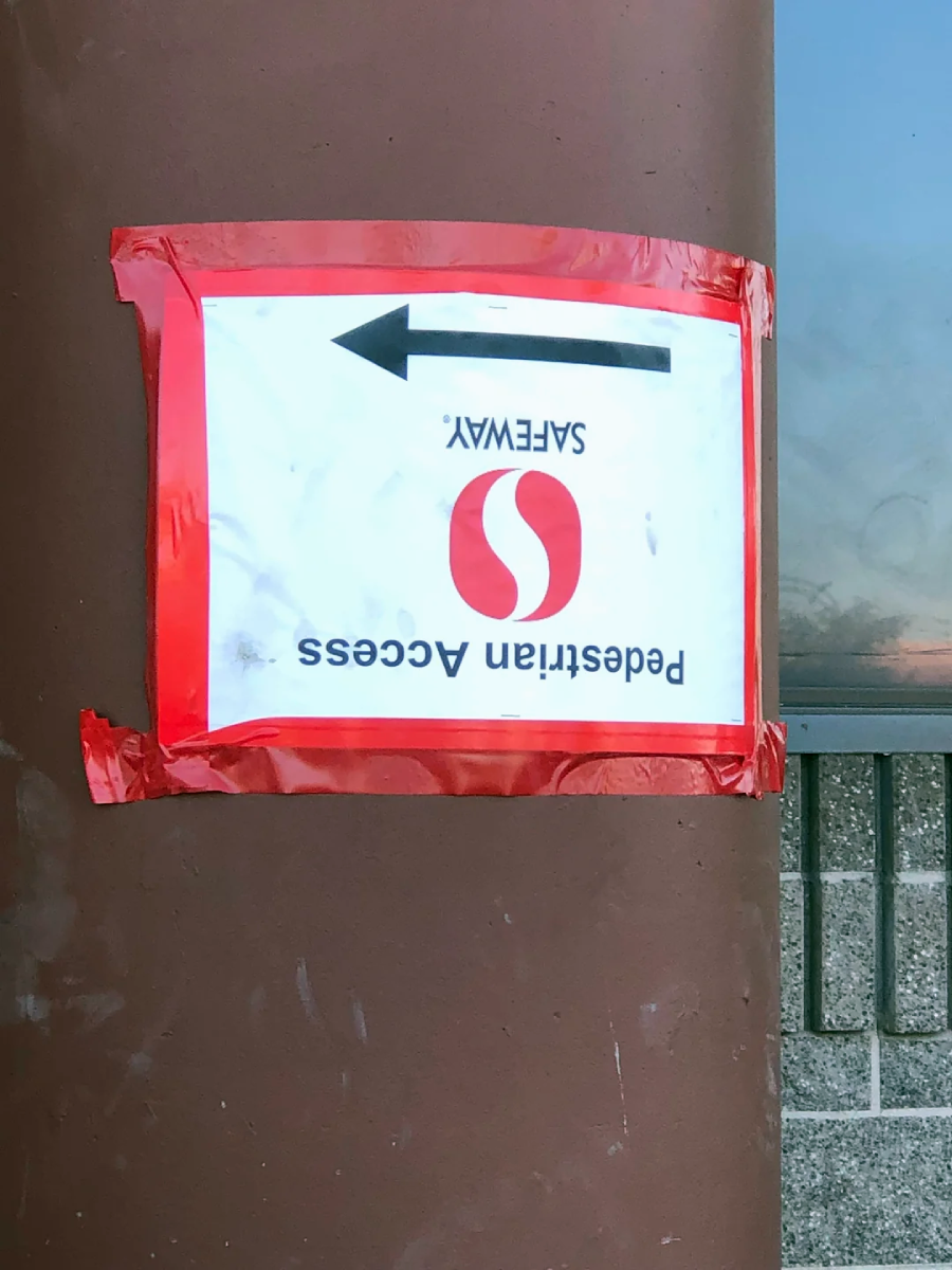 A sign taped to a brown pole with red tape reads “SAFeway Pedestrian Access” and an arrow, but the sign is upside down. The Safeway logo is also visible on the sign.