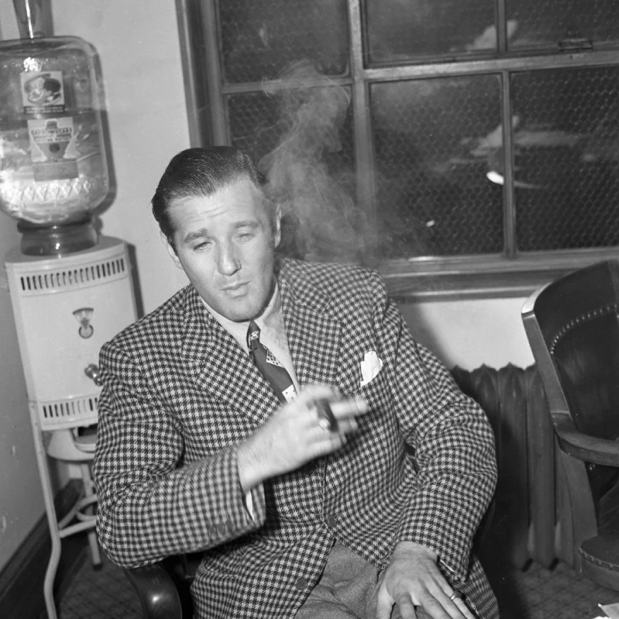 A man in a checkered suit sits indoors, holding a cigar with smoke rising. He is next to a water cooler and in front of a window, looking relaxed with his eyes partially closed.