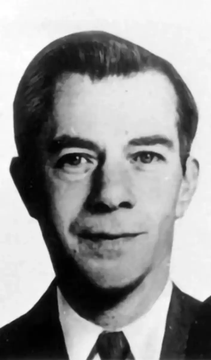 Black and white portrait of a man with short dark hair, wearing a suit and tie, looking directly at the camera with a neutral expression.