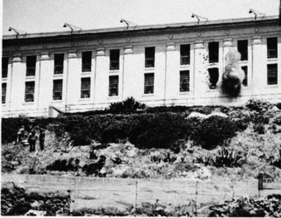 A black-and-white photo shows the exterior of a large building with tall windows, one of which is damaged by an explosion. Several people stand on the ground outside among bushes.