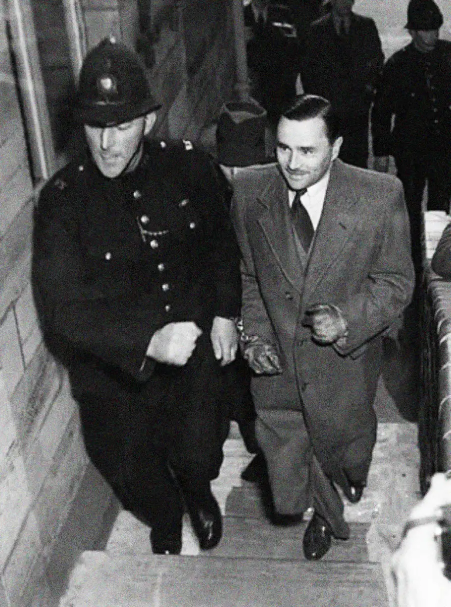 A police officer escorts a handcuffed man in a suit and overcoat up a set of stairs, with other people and police visible in the background. The scene appears to be from a past era.