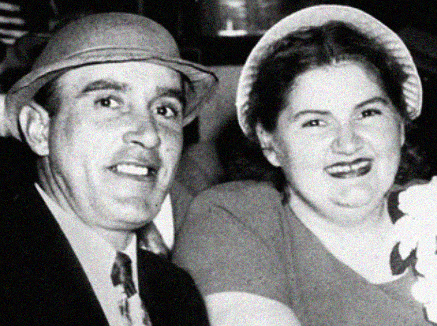 A smiling man in a suit and hat sits next to a smiling woman wearing a dress and a light-colored hat. The photo is black and white and appears to be vintage.