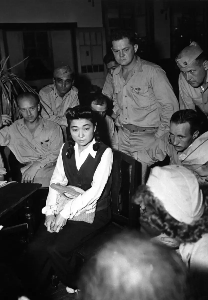 A Japanese woman sits at a table holding a woven bag, surrounded by several men in military uniforms, who are closely watching her in a crowded room.