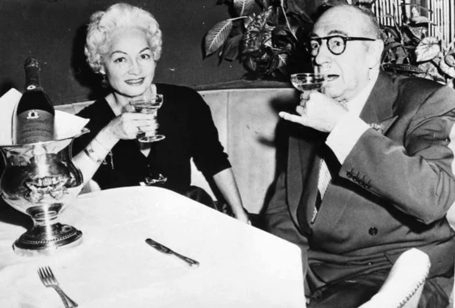 An older woman and man sit at a restaurant table, smiling and raising glasses of champagne. A bottle in an ice bucket and cutlery are on the white-tableclothed table. The background features plants and a dark wall.