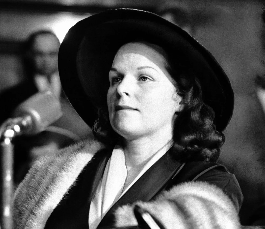 A woman in a wide-brimmed hat and fur-trimmed coat speaks into a vintage microphone, with an audience blurred in the background. The image is in black and white.