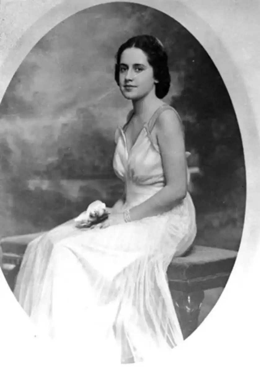 A young woman in a light-colored, sleeveless gown sits on a bench, holding a small object in her hand. The photo is black and white with a soft, oval vignette and a studio-style background.
