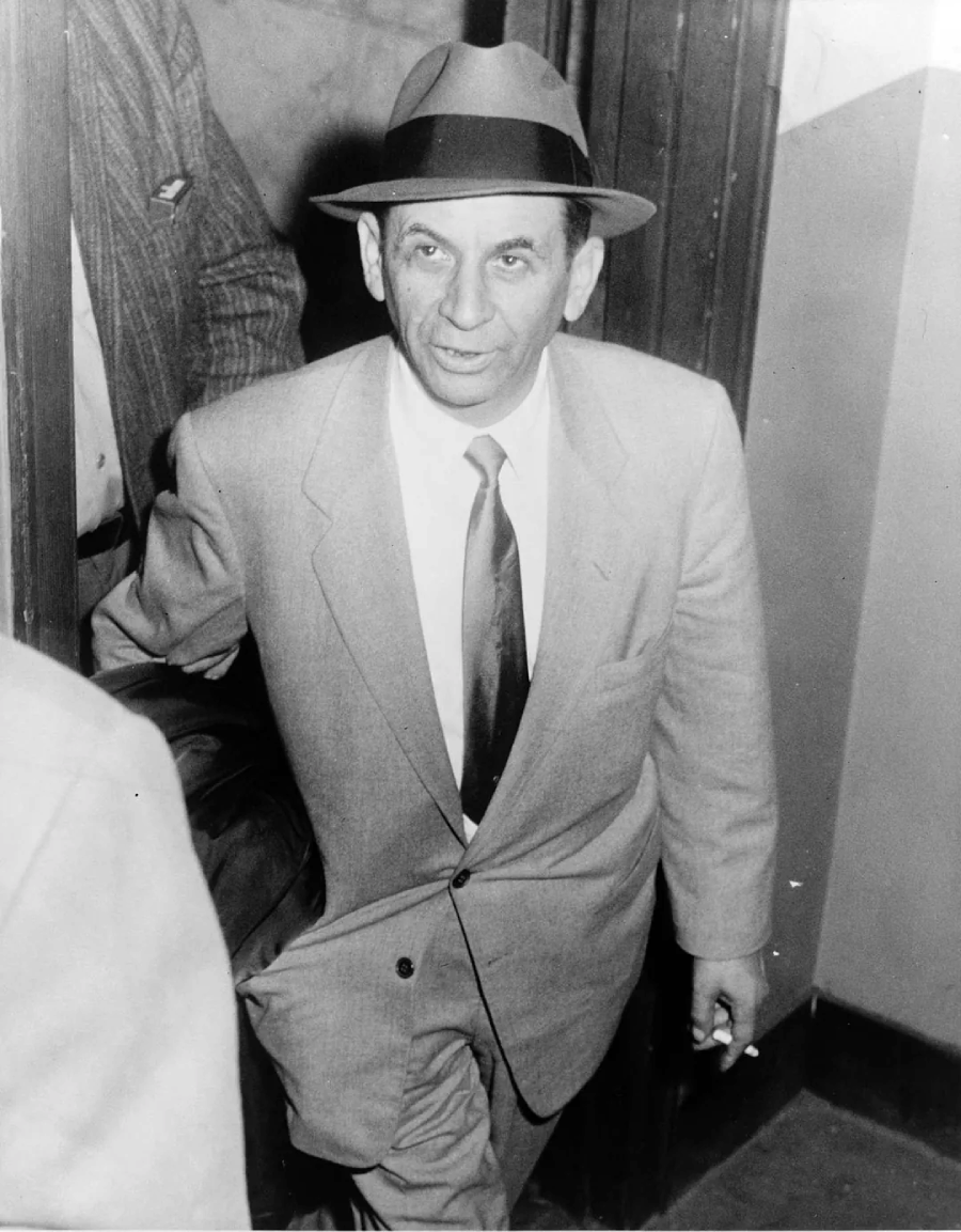 A man in a suit, tie, and fedora hat walks through a doorway, looking slightly upward. He holds a cigarette in his right hand and appears to be mid-step, with other people partially visible around him. The photo is black and white.