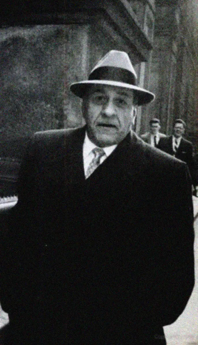A man in a suit and tie wearing a fedora hat stands outside on a city street, looking at the camera. The image is in black and white and has a vintage appearance. Two men are walking in the background.