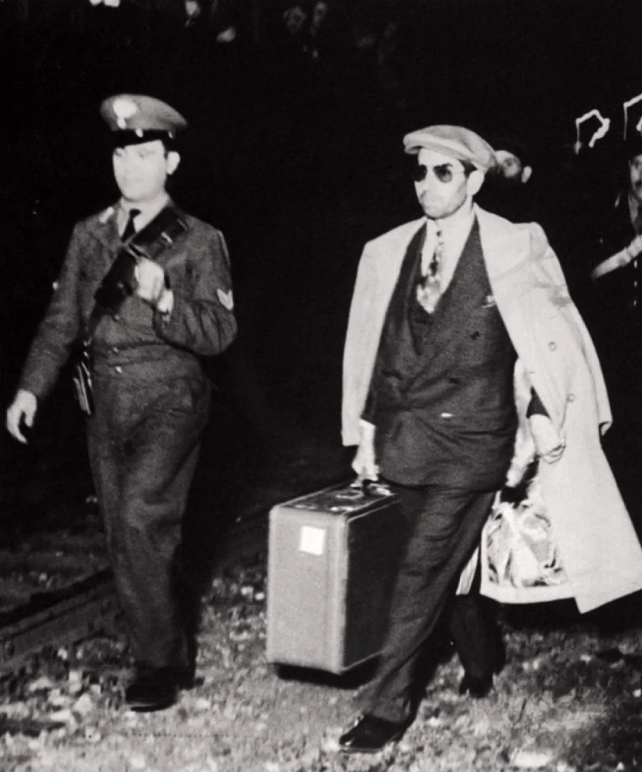 A man in a suit, sunglasses, and a hat walks alongside a uniformed officer. The man carries a suitcase and has a coat draped over his shoulders. They appear to be outdoors at night.