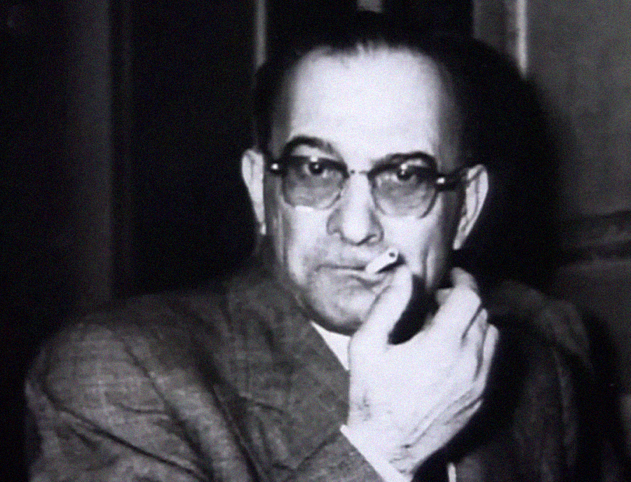 Black and white photo of a middle-aged man wearing glasses and a suit, holding a cigarette near his mouth, looking directly at the camera with a serious expression.