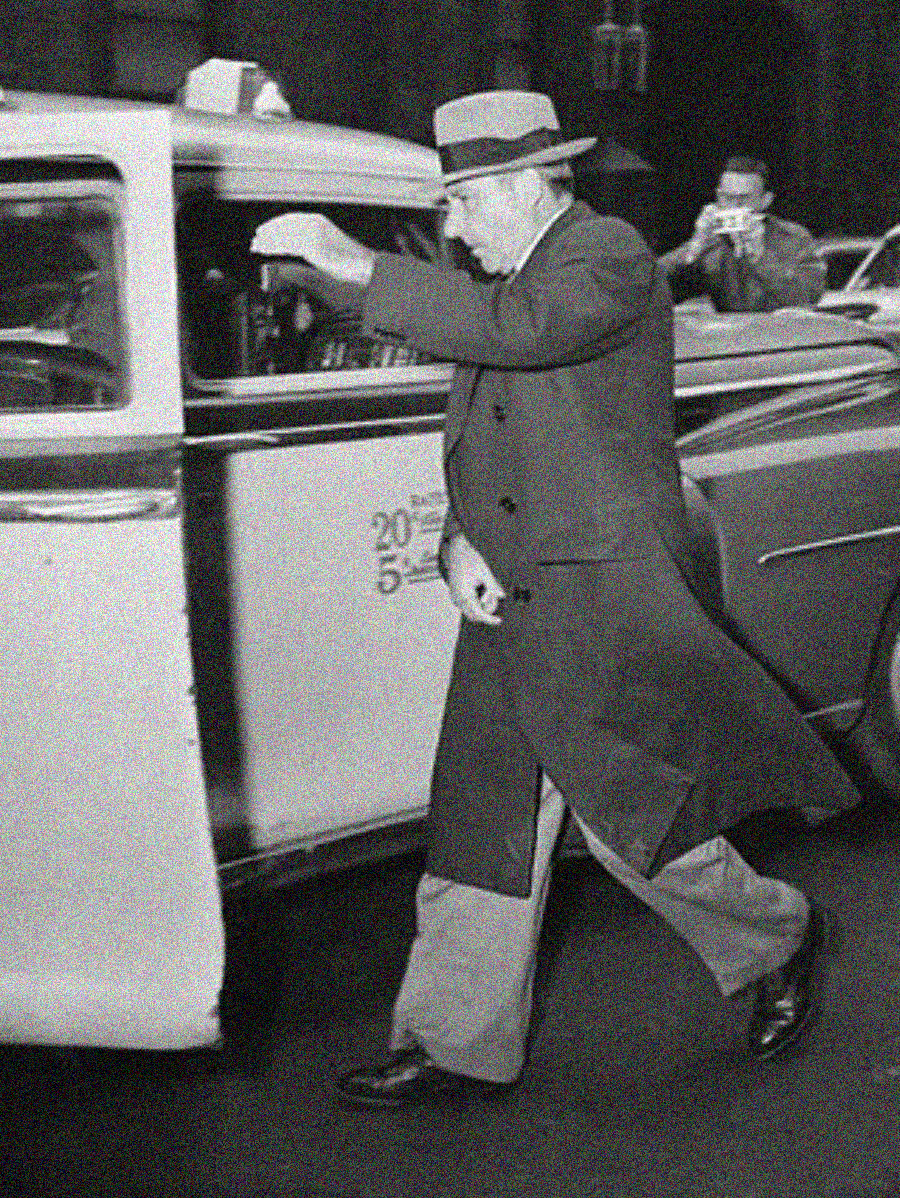 A man in a long coat and hat quickly steps toward a taxi with its door open, shielding his face from the camera. Another person in the background is taking a photo. The image is in black and white.