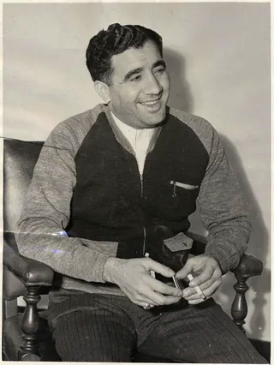A man with dark, wavy hair sits on a wooden chair, smiling, wearing a two-tone jacket and pinstriped pants. He holds a cigarette in one hand and a lighter in the other. The photo is black and white.