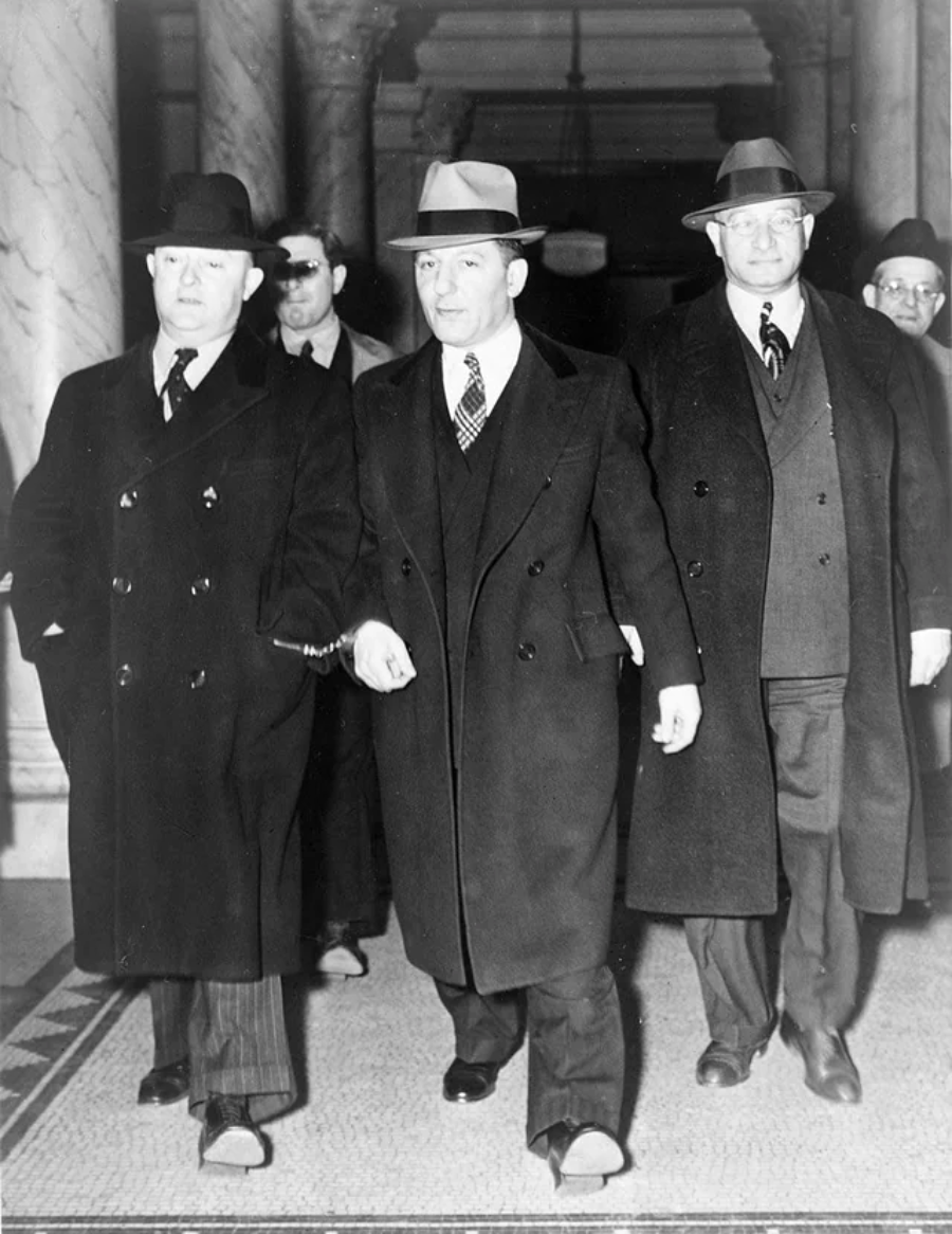 Three men in dark overcoats and hats walk side by side in a marble hallway, followed by two other men. The central figure strides confidently, and all appear serious and formal, dressed in suits and ties.