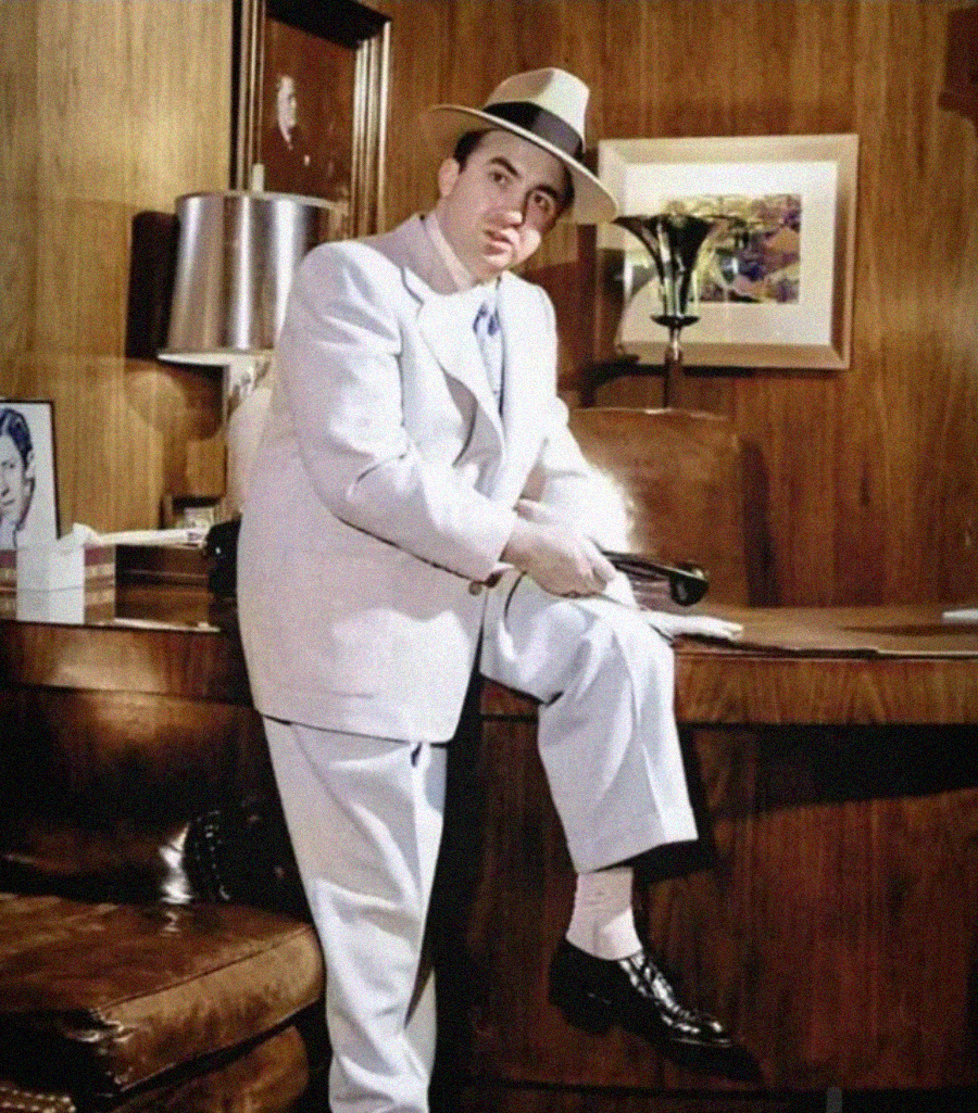 A man in a light-colored suit and hat leans against a wooden desk in a vintage office, resting one foot on a chair, with framed photos and a lamp visible in the background.