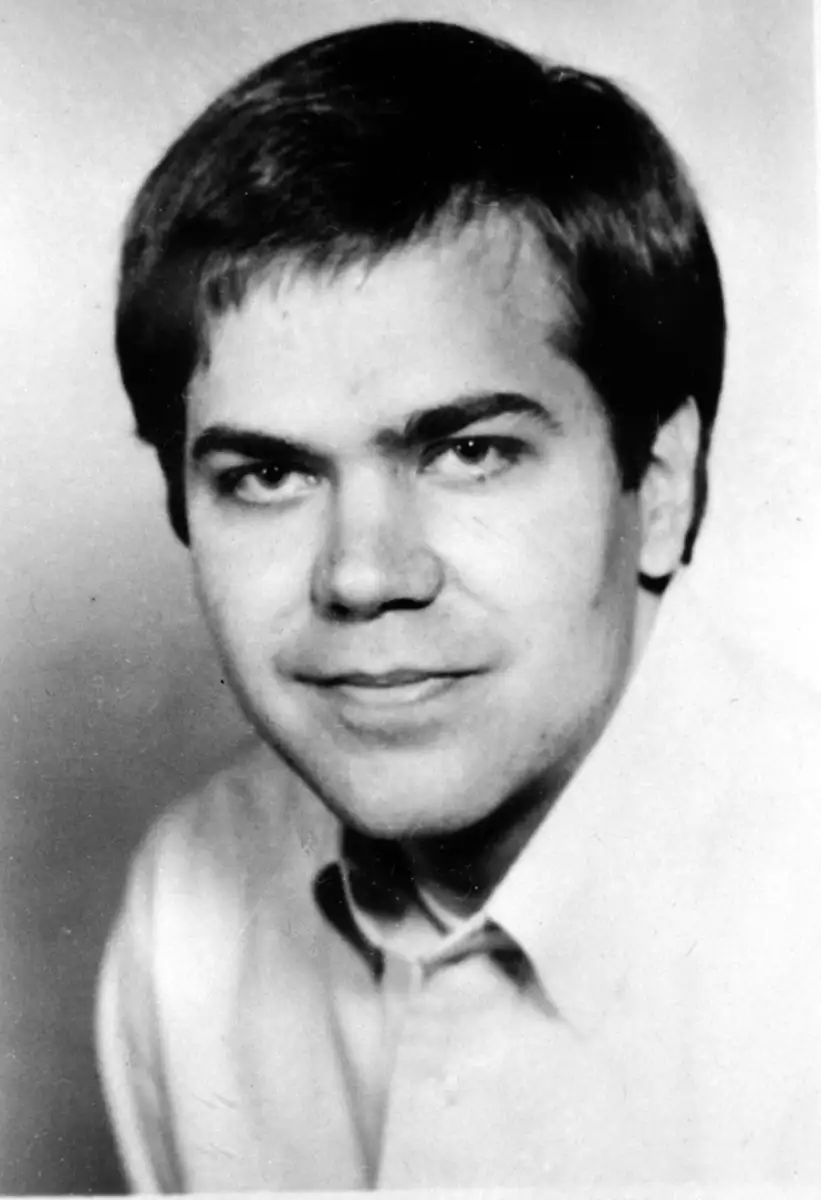 A black-and-white portrait of a young man with short dark hair, wearing a light-colored collared shirt, looking slightly past the camera with a neutral expression.
