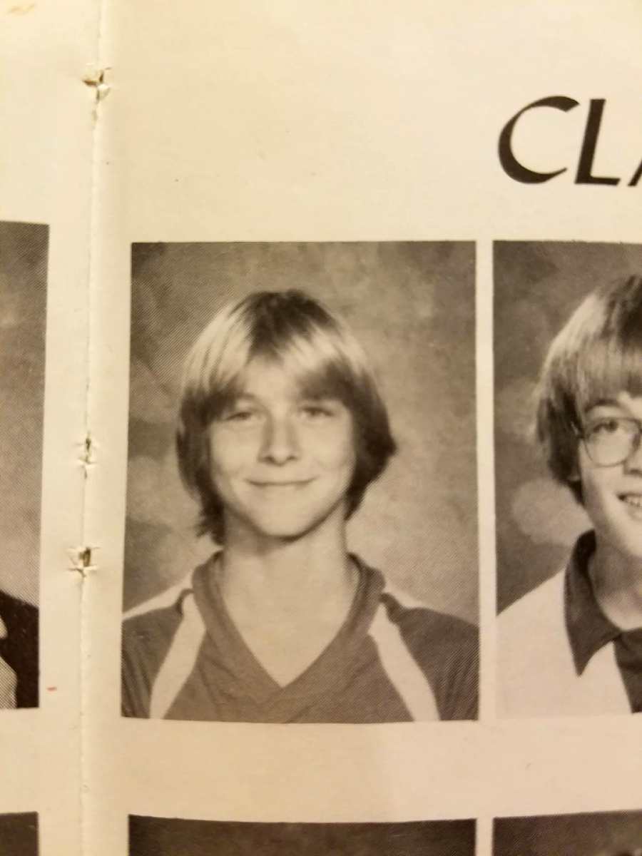 A black and white yearbook photo of a smiling young person with medium-length, light hair, wearing a collared shirt. There is part of another portrait visible on each side.