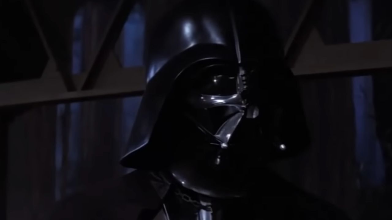 A person wearing a shiny black helmet and mask stands in a dimly lit room with shadows and blue light in the background.