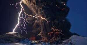 A dramatic volcanic eruption at night shows thick, dark ash clouds, bright orange lava, and several vivid bolts of lightning striking down, with snow-covered ground in the foreground.