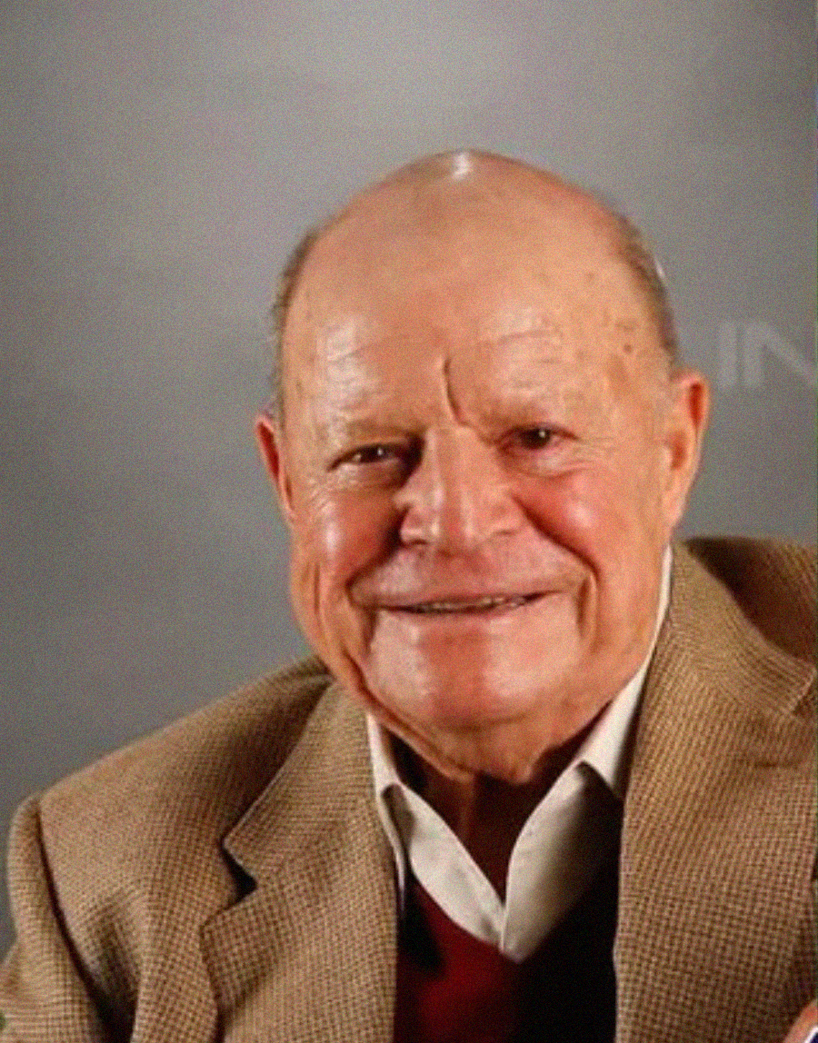 An older man with a bald head and light complexion smiles at the camera. He is wearing a tan blazer over a cream shirt and red sweater, set against a plain gray background.