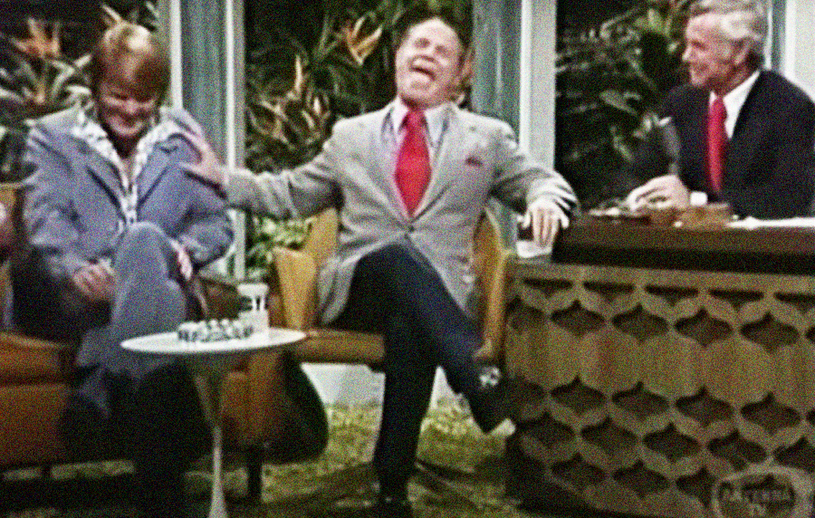Three men in suits sit and laugh together on a vintage talk show set, with one host at a desk and two guests seated on chairs, surrounded by retro decor and plants in the background.