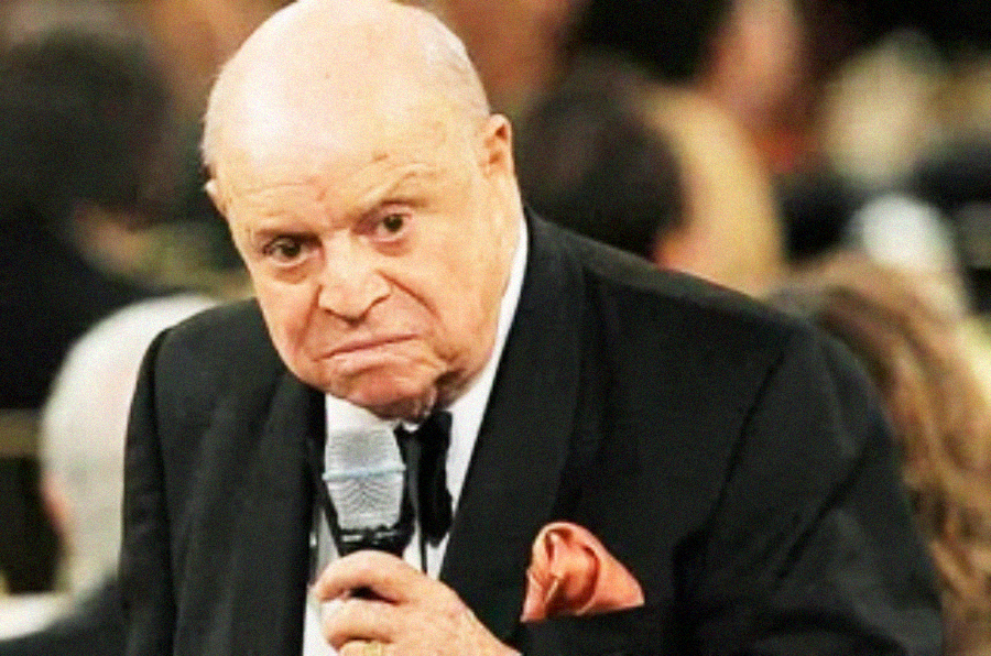 An older man in a tuxedo with a pink pocket square holds a microphone and looks forward with a serious, slightly frowning expression. The background is blurred, showing several people.