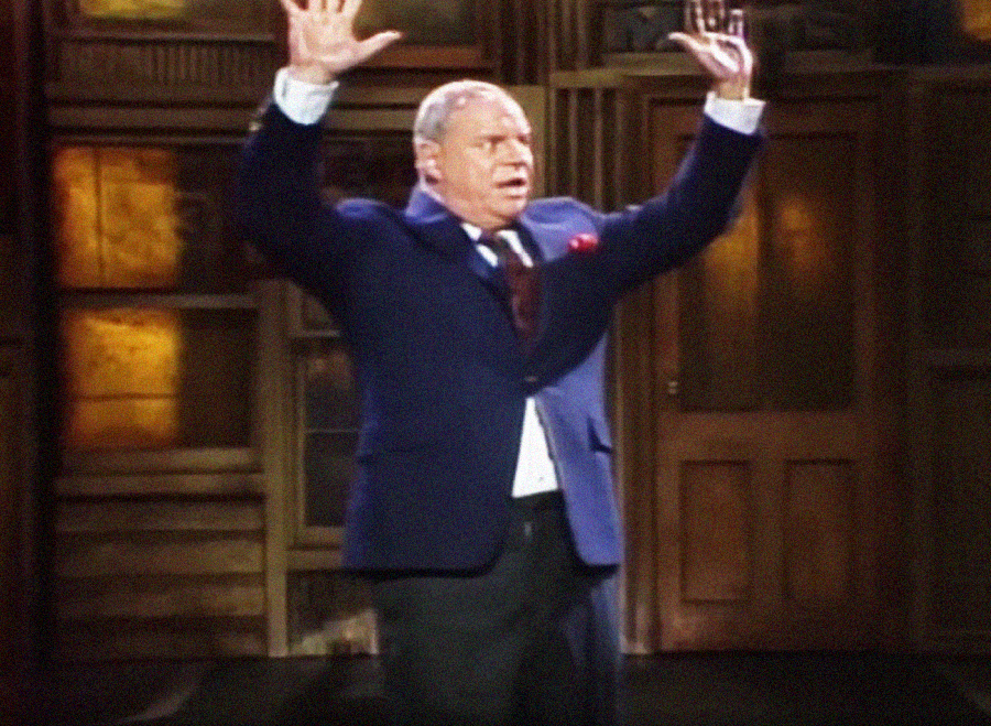 A man in a dark suit stands indoors with both arms raised, palms open, and an expressive facial expression. Wooden doors and panels are visible in the background.