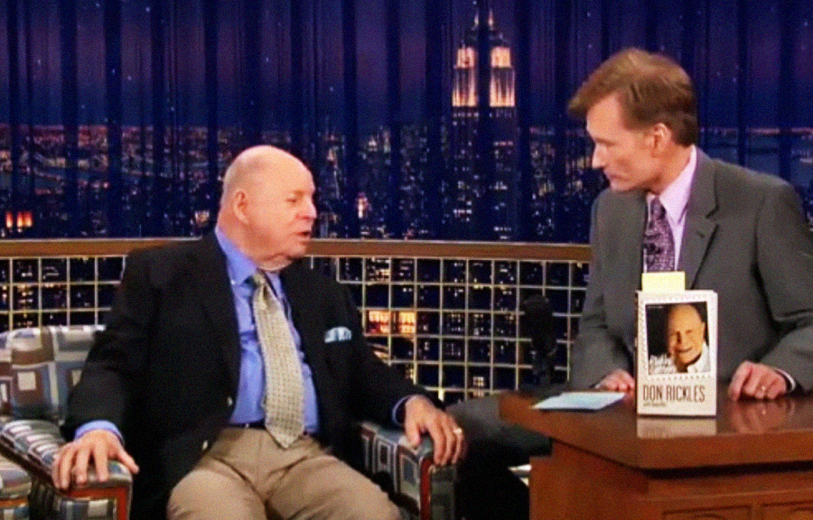 An older man in a suit sits and talks with a younger man in a suit on a late-night talk show set. A book with the older man's photo and name is displayed on the desk between them.