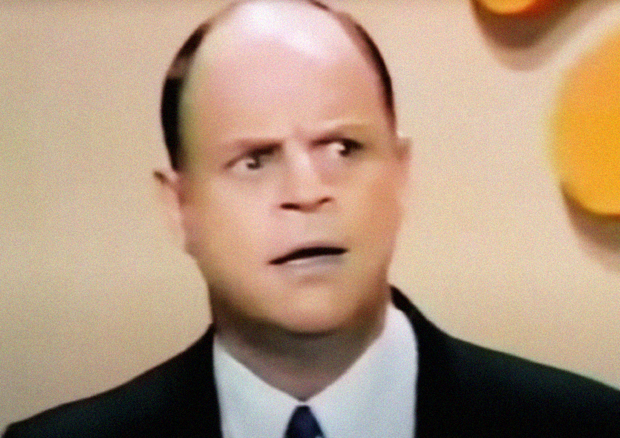 A man in a suit and tie looks surprised or confused, with raised eyebrows and a slightly open mouth, standing in front of a beige background with yellow shapes.