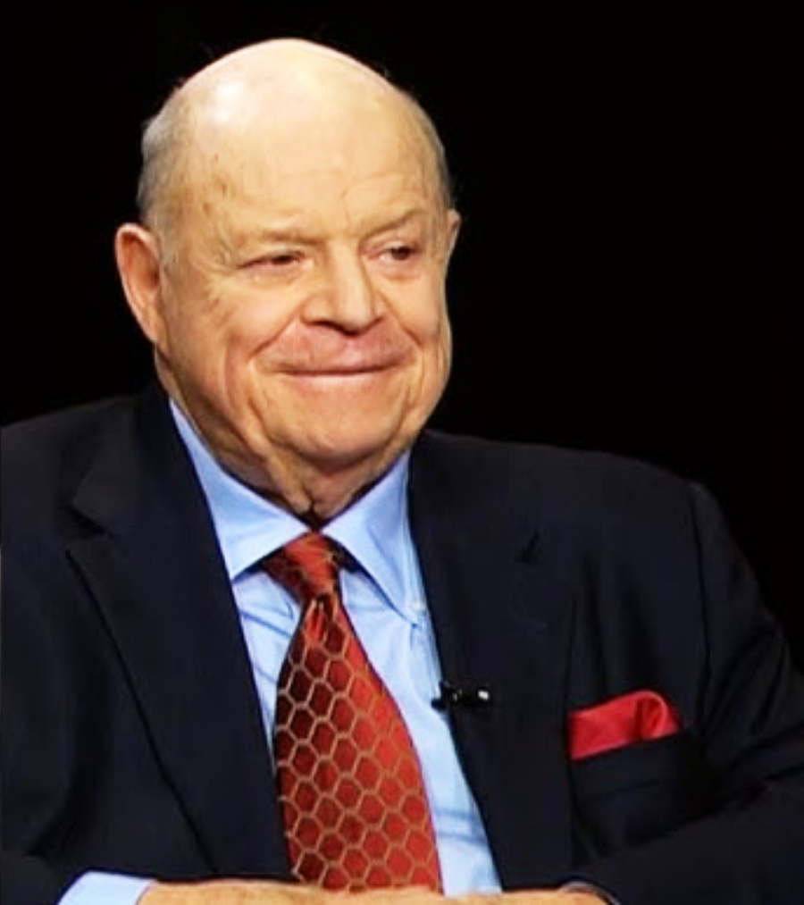 An older man with a bald head smiles while wearing a dark suit, light blue shirt, patterned red tie, and red pocket square, seated against a black background.