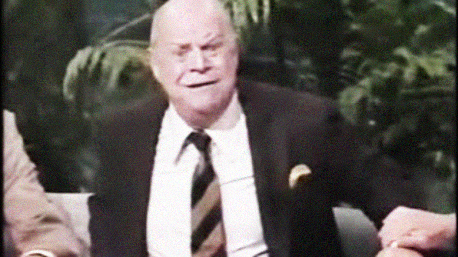 An older man in a suit and striped tie sits on a couch, smiling with a slightly raised eyebrow, against a background of green plants.