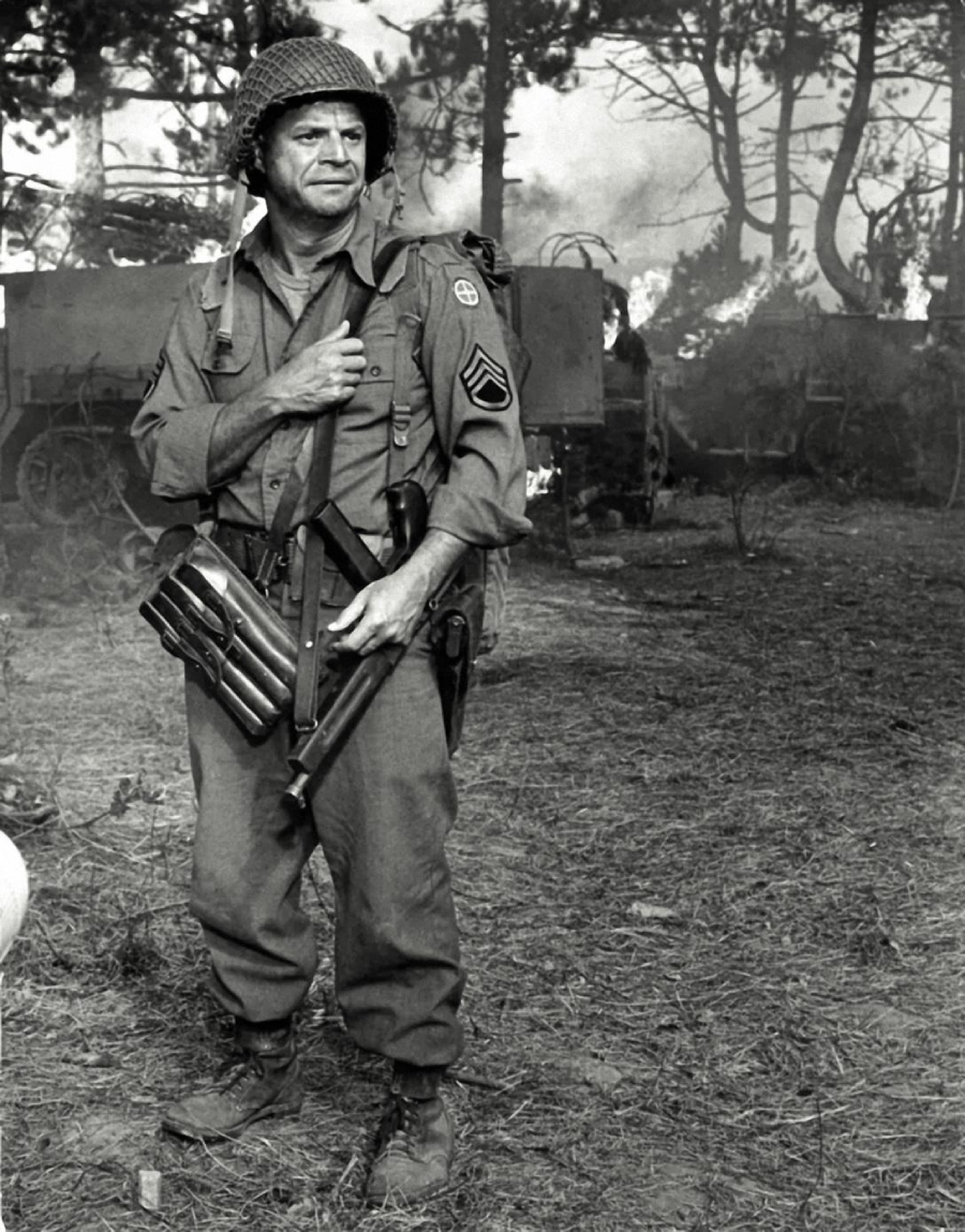A World War II soldier in uniform stands outdoors, holding a submachine gun and several grenades, with smoke and military vehicles in the background.