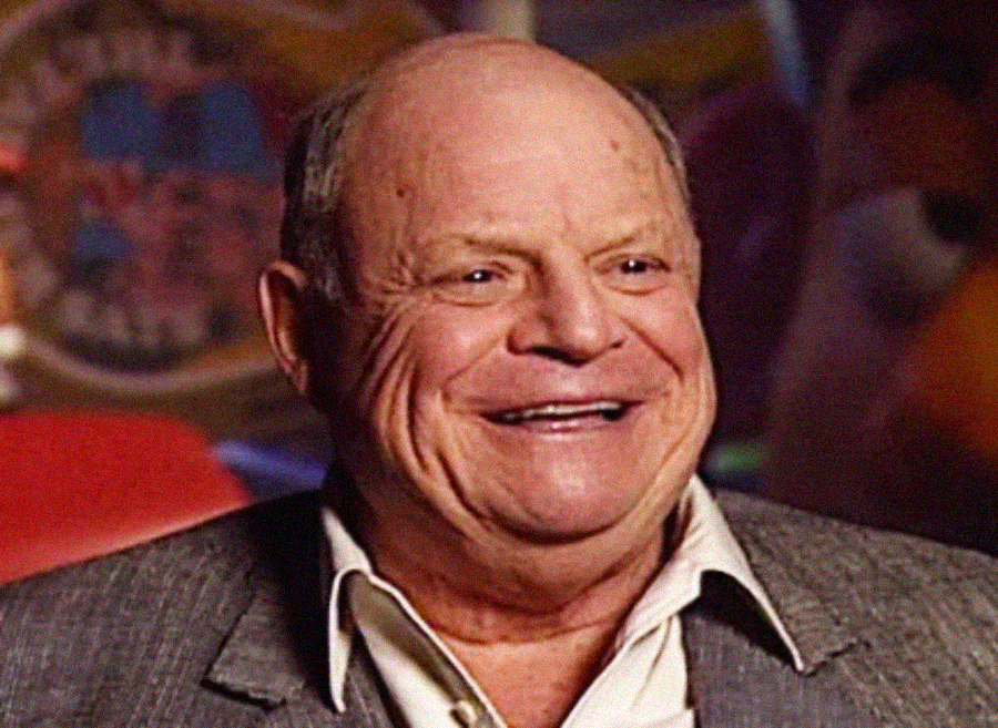 An older man with a bald head and a big smile wears a gray jacket and white shirt, sitting indoors with colorful, blurred shapes in the background.
