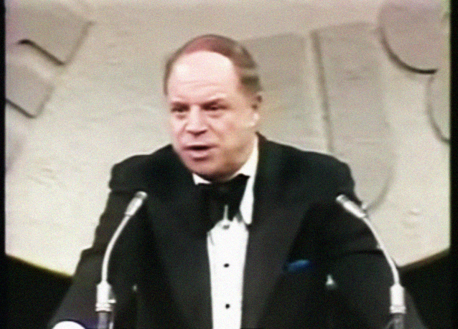A man in a tuxedo stands at a podium with two microphones, speaking. The background features large abstract shapes and muted colors.