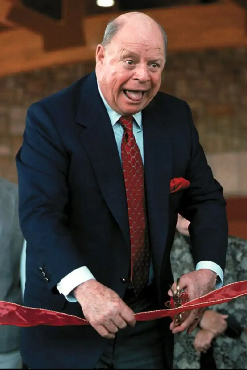 An older man in a dark suit and red tie smiles widely while cutting a red ribbon with large scissors at an indoor ribbon-cutting ceremony.