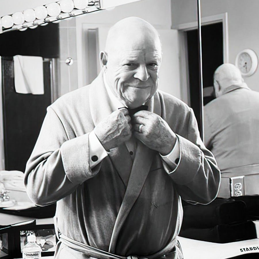 An older man in a light-colored robe adjusts his collar and smiles in front of a mirror in a well-lit dressing room, with his reflection visible behind him.
