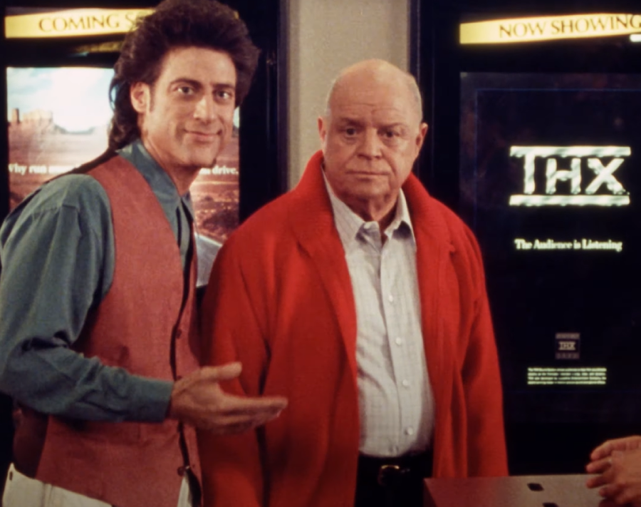 Two men stand indoors in front of movie posters. One man wears a red vest and blue shirt, smiling and gesturing, while the other man wears a red jacket and looks serious.